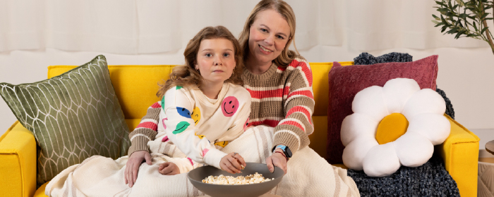 Parent and child sit on a yellow couch sharing a bowl of popcorn, illustrating calm, connected moments that support emotional wellness and positive routines at home.