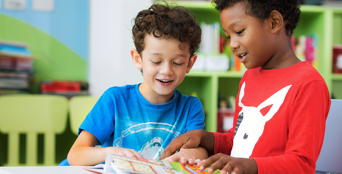 Two young boys smiling and reading a book together in a brightly colored classroom or library, promoting peer connection and early childhood literacy."