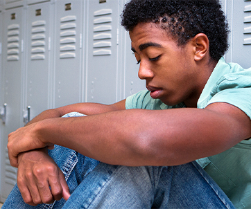 Teen boy sitting on the floor with eyes closed and arms around his knees in front of school lockers, representing youth mental health challenges highlighted in CDC surveys.
