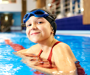 Smiling girl in a swimsuit and swim cap rests at the edge of a pool, promoting physical activity as a micro-habit to support mental well-being in kids.