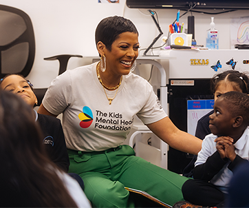 Tamron Hall, wearing a Kids Mental Health Foundation shirt, smiles and engages with a group of young students during a classroom visit, promoting mental health awareness and connection.