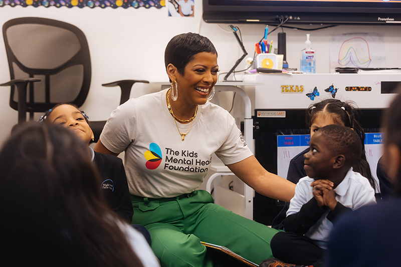 Tamron Hall visits a classroom, engaging with young students while representing The Kids Mental Health Foundation. She smiles warmly, wearing a branded foundation t-shirt, as she interacts with children in a positive and supportive learning environment. The visit highlights her advocacy for children's mental health awareness and education.